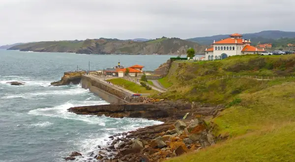 Costa de San Vicente de la Barquera a Comillas con edificios blancos y tejados naranjas, mar agitado y acantilados verdes.