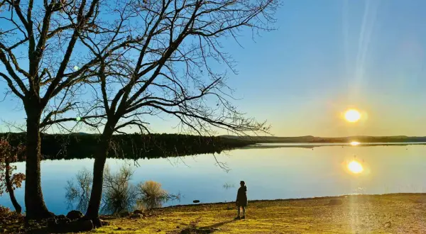 Una persona contempla el atardecer en la playa de Soria, con árboles desnudos y el sol reflejado en el agua serena.