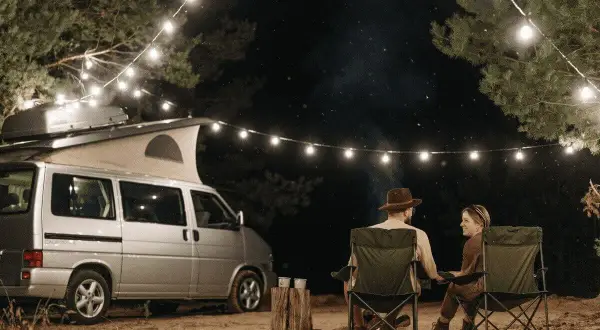 Pareja acampando bajo luces de feria en la costa atlántica francesa. Una furgoneta camper y sillas plegables crean un ambiente acogedor.