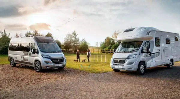 Dos vehículos, un camper y una caravana, estacionados al aire libre. Dos mujeres y sus perros disfrutan de un día soleado.