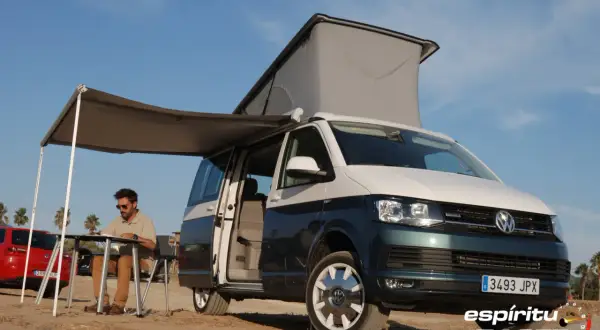 Hombre sentado en mesa plegable bajo toldo de camper en playa, con medidas california beach.