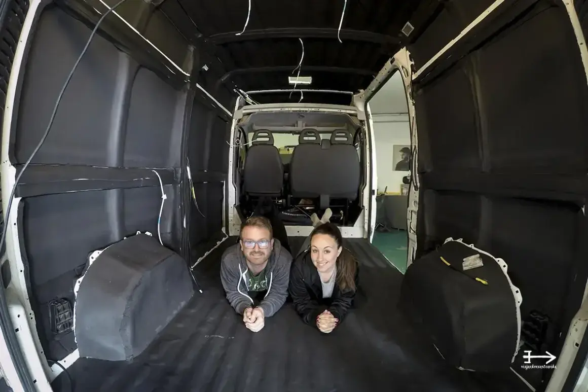 Pareja sonríe tumbada en el suelo de una furgoneta camper en proceso de aislamiento. Merece la pena aislar furgoneta camper para futuras aventuras.