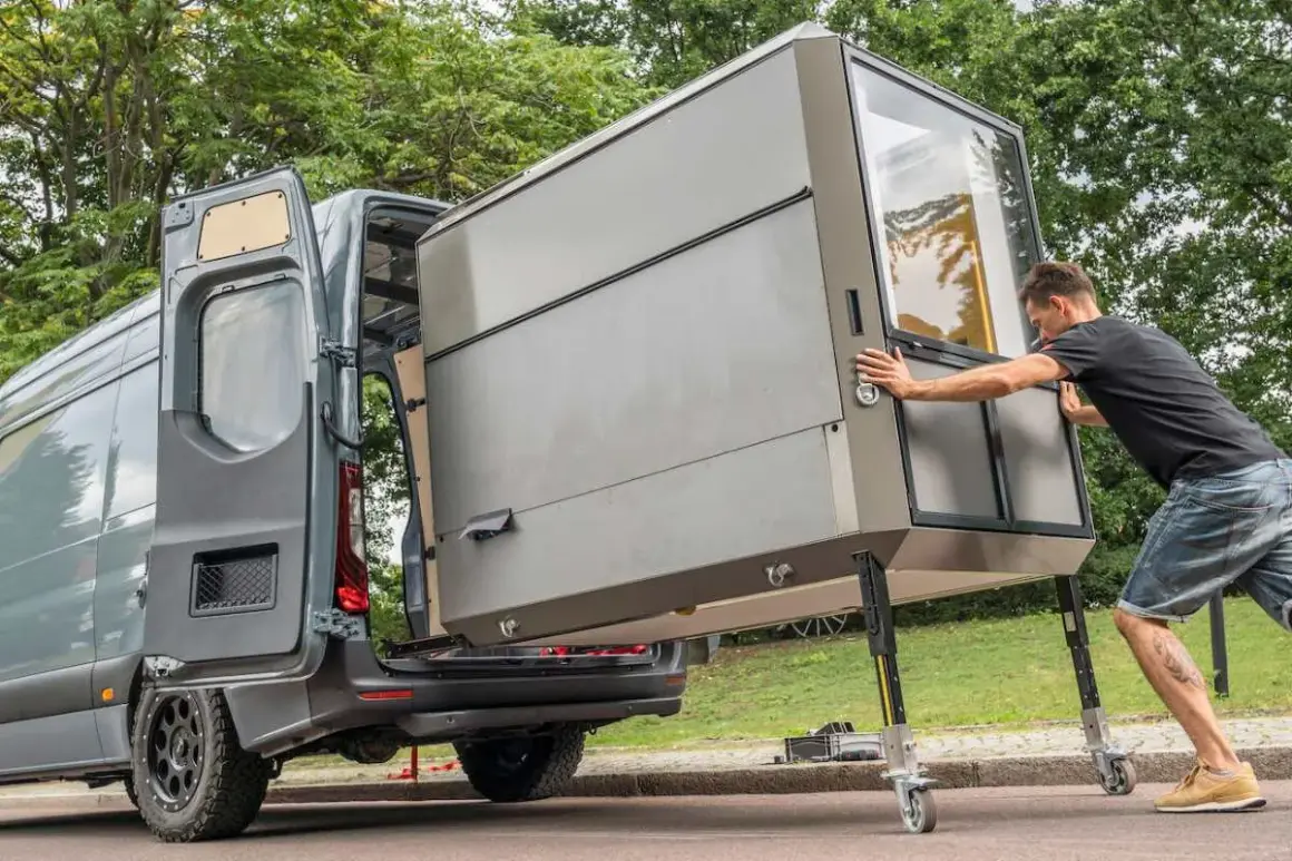 Hombre empuja módulo habitacional con ruedas hacia una furgoneta. ¡Es un sistema plug and play para tu aventura!