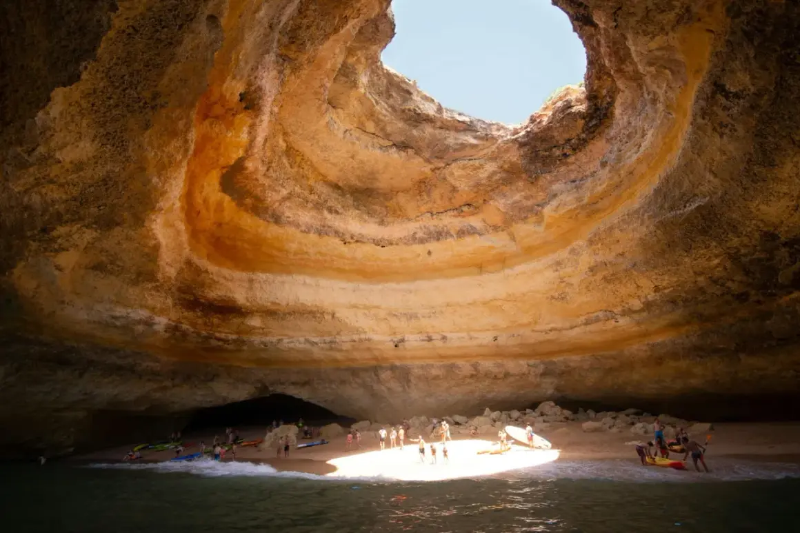 Interior de una cueva marina con un agujero en el techo, donde está Benagil. Gente en kayaks y en la playa disfrutan del sol.