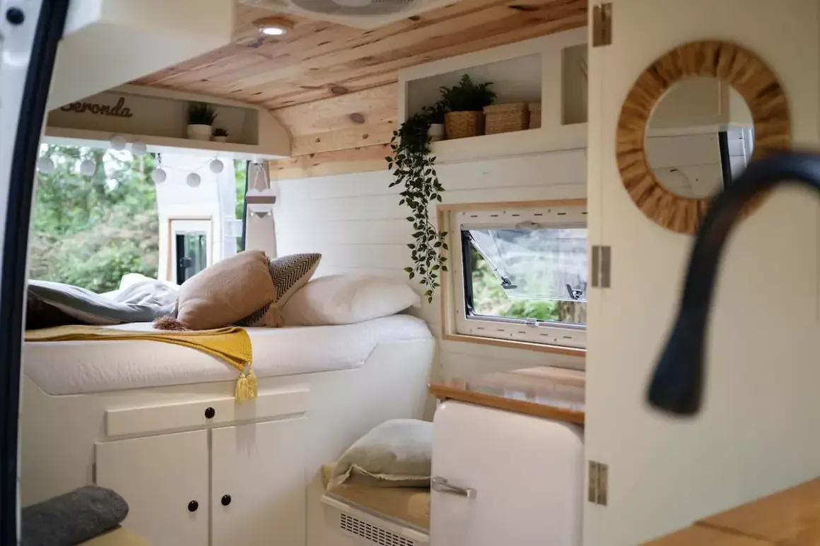 Interior de una camper con cama acogedora, cojines y mantas. Muestra cómo llevar la ropa de forma organizada en este espacio.