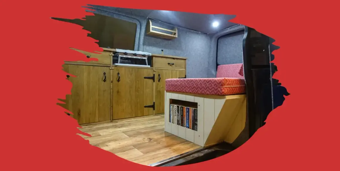 Interior de caravana con cocina, cama y estanterías. El suelo de madera renovado da un aspecto acogedor.
