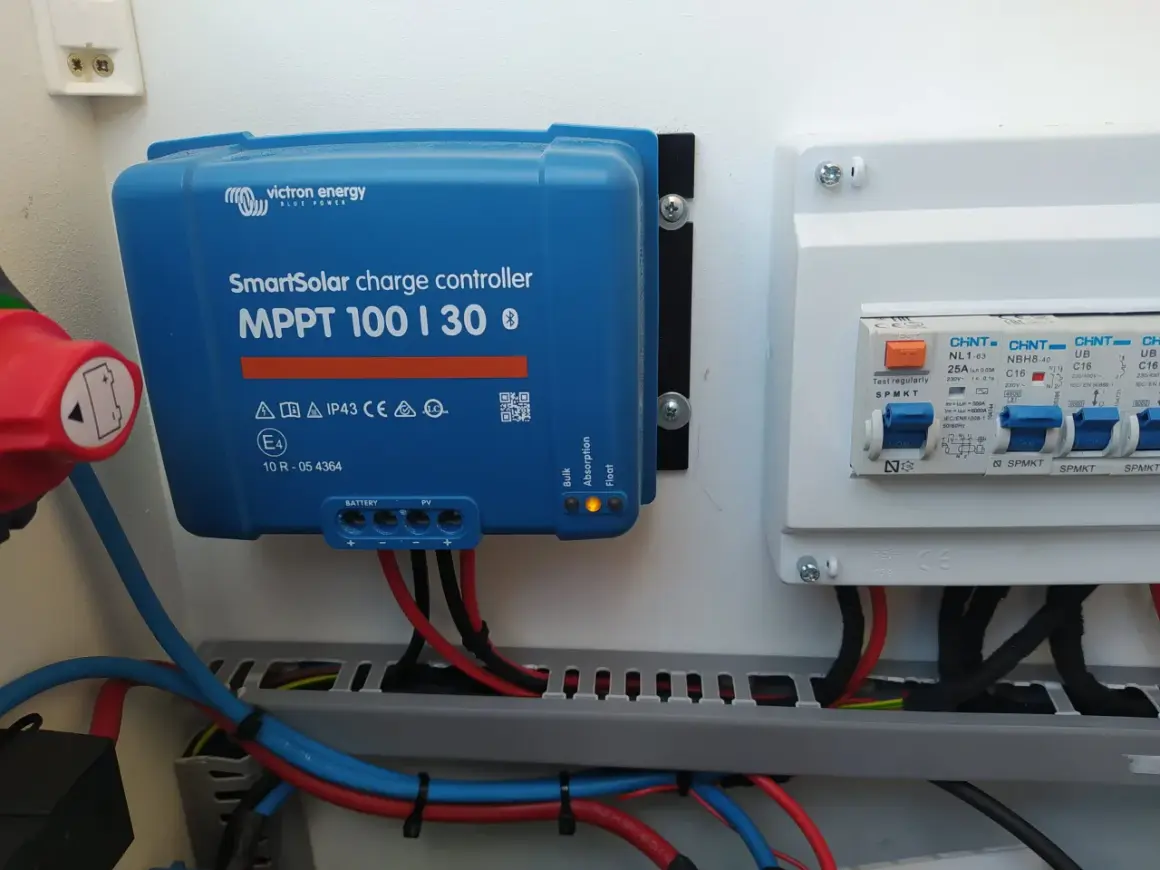 Controlador de carga solar Victron MPPT 100|30, con luces indicadoras de estado y cables conectados. A su derecha, un cuadro eléctrico con interruptores.
