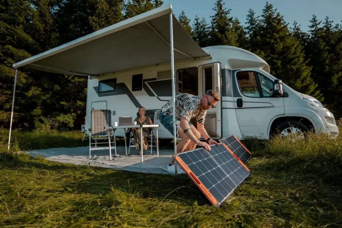 Hombre instala paneles solares, quizás en serie o paralelo, junto a una autocaravana. Mujer trabaja en portátil bajo toldo.