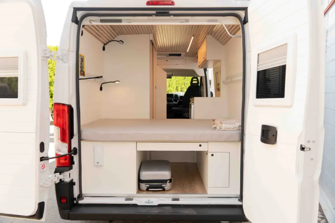 Interior de una camper con cama, almacenamiento y un pequeño equipaje. La altura camper permite aprovechar cada rincón.