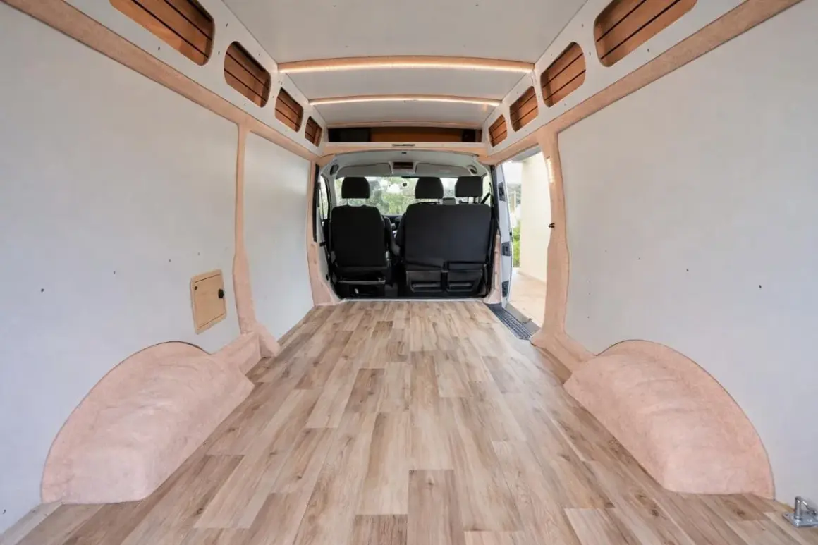 Interior de furgoneta camper con suelo de madera. Las paredes están revestidas y hay asientos en la parte delantera.