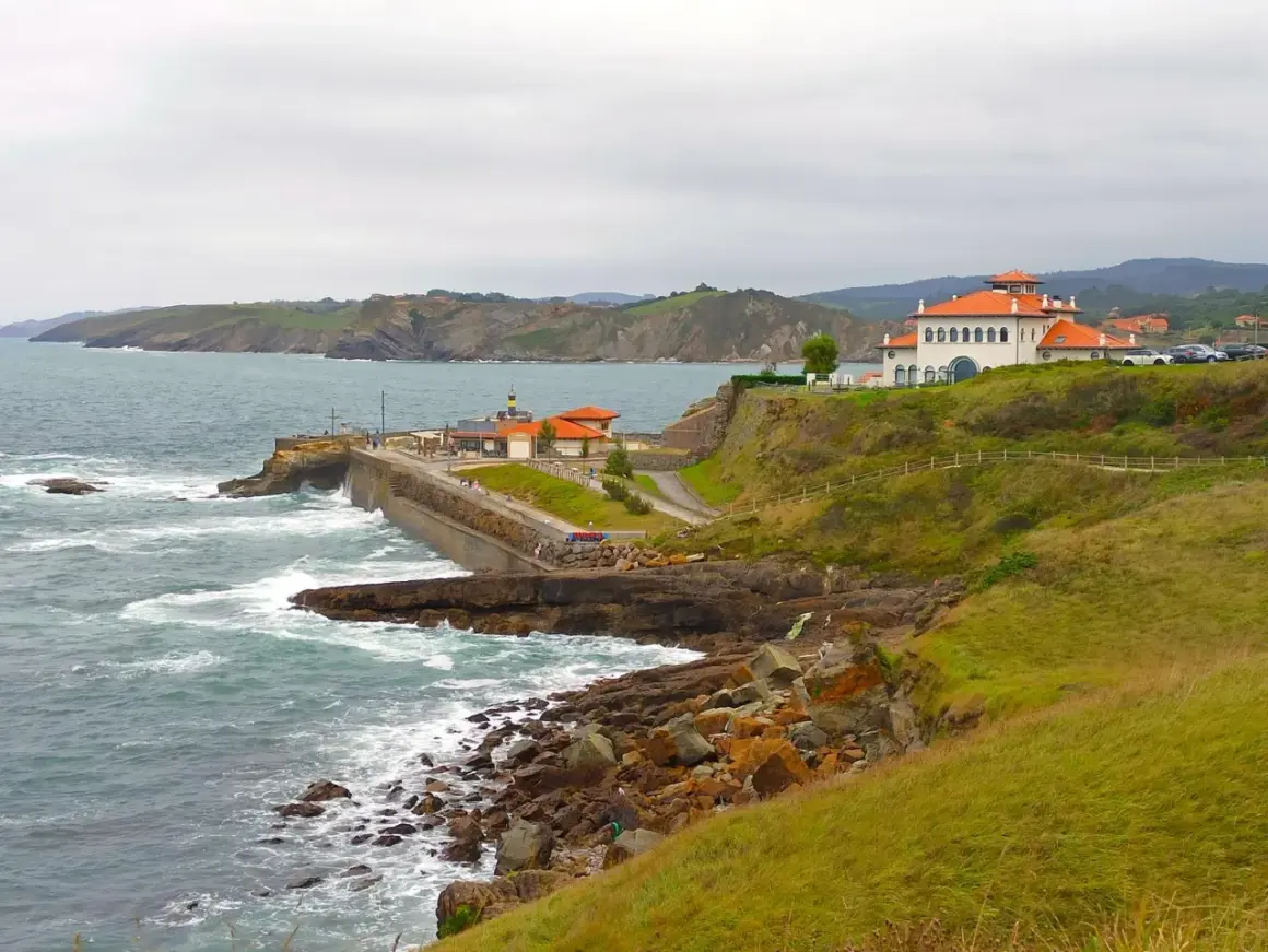 Costa de San Vicente de la Barquera a Comillas con edificios blancos y tejados naranjas, mar agitado y acantilados verdes.