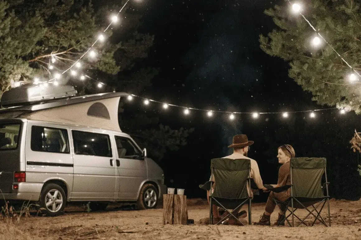 Pareja acampando bajo luces de feria en la costa atlántica francesa. Una furgoneta camper y sillas plegables crean un ambiente acogedor.