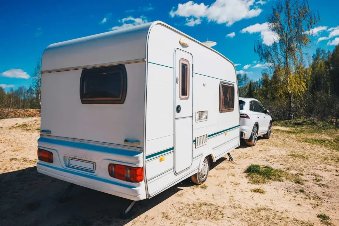 Caravana blanca lista para el transporte, aparcada junto a un coche en un día soleado.