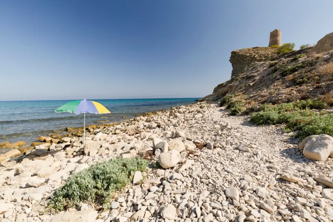 Sombrilla de colores en la playa de las covaticas, con mar turquesa y torre antigua en el acantilado.
