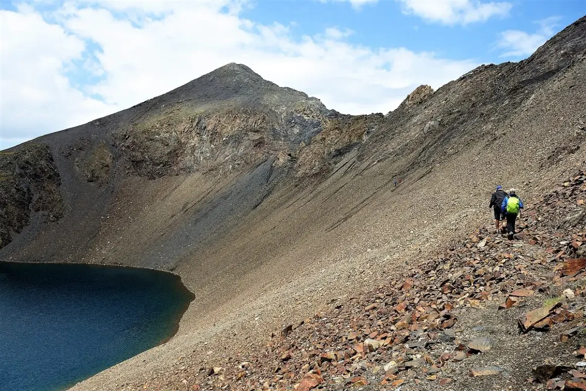 Senderistas ascienden por un sendero pedregoso junto a un lago azul, con la imponente silueta de los ibones de Bachimaña al fondo.