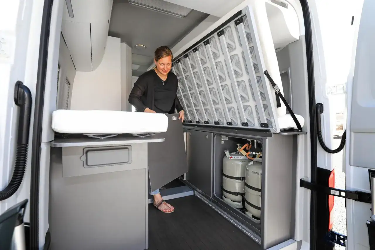 Interior de una Volkswagen California con ba&ntilde;o, mostrando la cama desplegada y los tanques de propano.