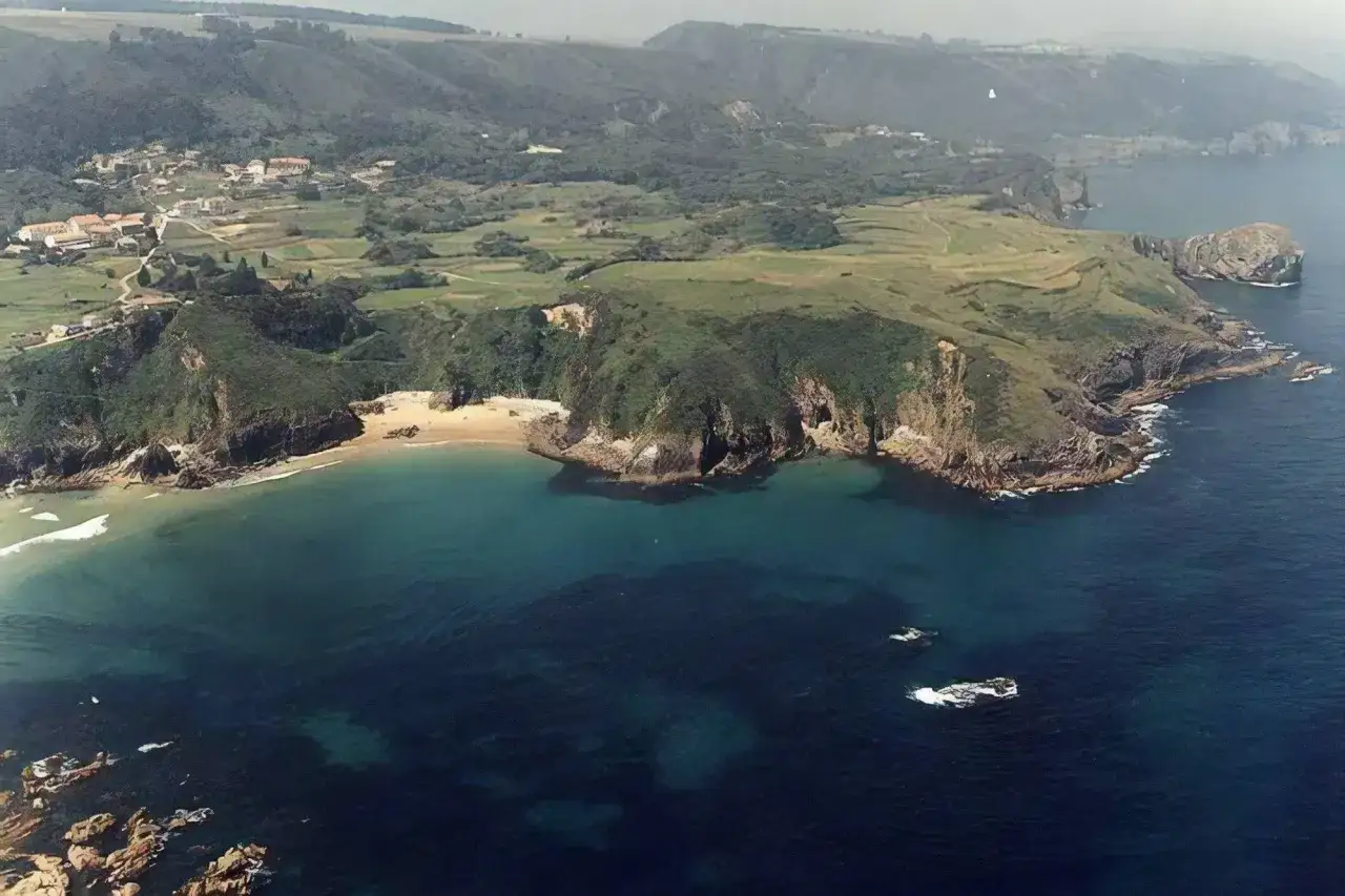 Vista a&eacute;rea de la playa de Aramal, con acantilados verdes, arena dorada y aguas turquesas. Un pueblo se asoma entre las colinas.