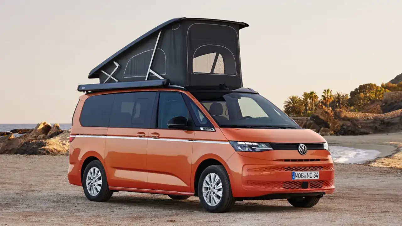 Furgoneta naranja con techo elevable en una playa de arena, lista para aventuras con las medidas perfectas para explorar la costa de California.