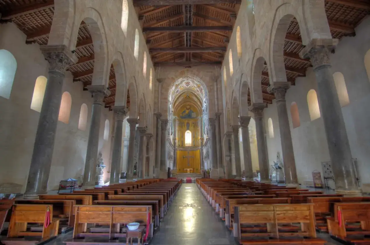 Interior de la catedral de Cefal&uacute;, con filas de bancos de madera, columnas imponentes y un mosaico bizantino en el &aacute;bside.