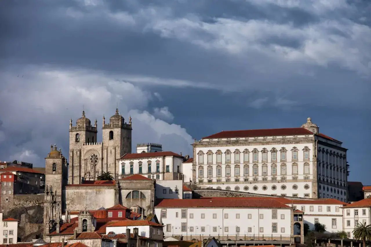 Edificios hist&oacute;ricos de Oporto bajo un cielo amenazante, con la catedral y el palacio a la vista. El oporto con lluvia se intuye en las nubes oscuras.