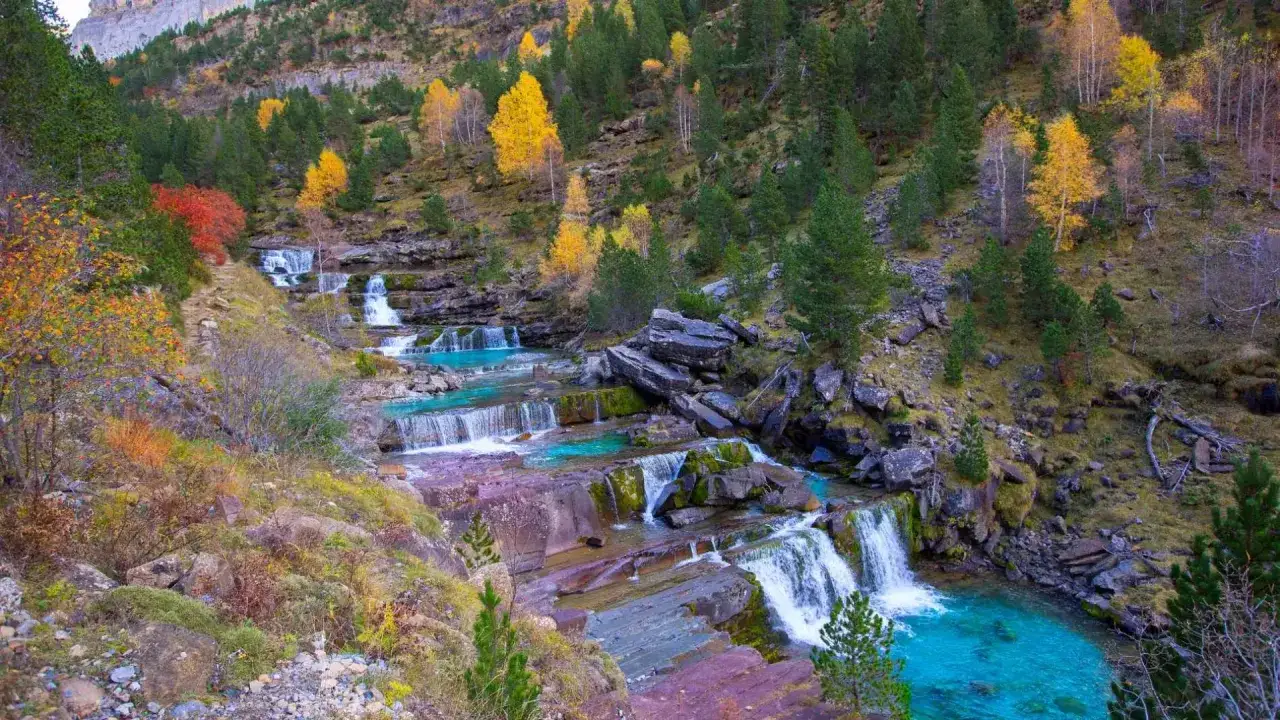Cascada en las gradas de soaso, con aguas turquesas y rocas rojizas, rodeada de árboles otoñales y vegetación verde.