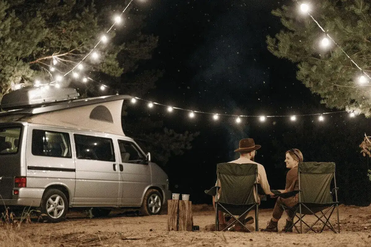 Pareja acampando bajo luces de feria en la costa atl&aacute;ntica francesa. Una furgoneta camper y sillas plegables crean un ambiente acogedor.