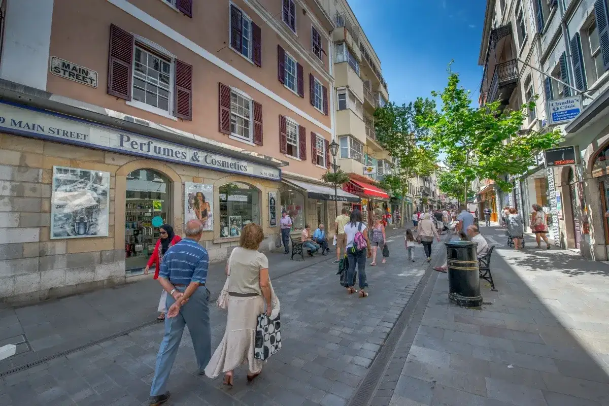 Gente paseando por la bulliciosa Gibraltar altstadt, con tiendas de perfumes y cosm&eacute;ticos, y edificios hist&oacute;ricos bajo un cielo azul.