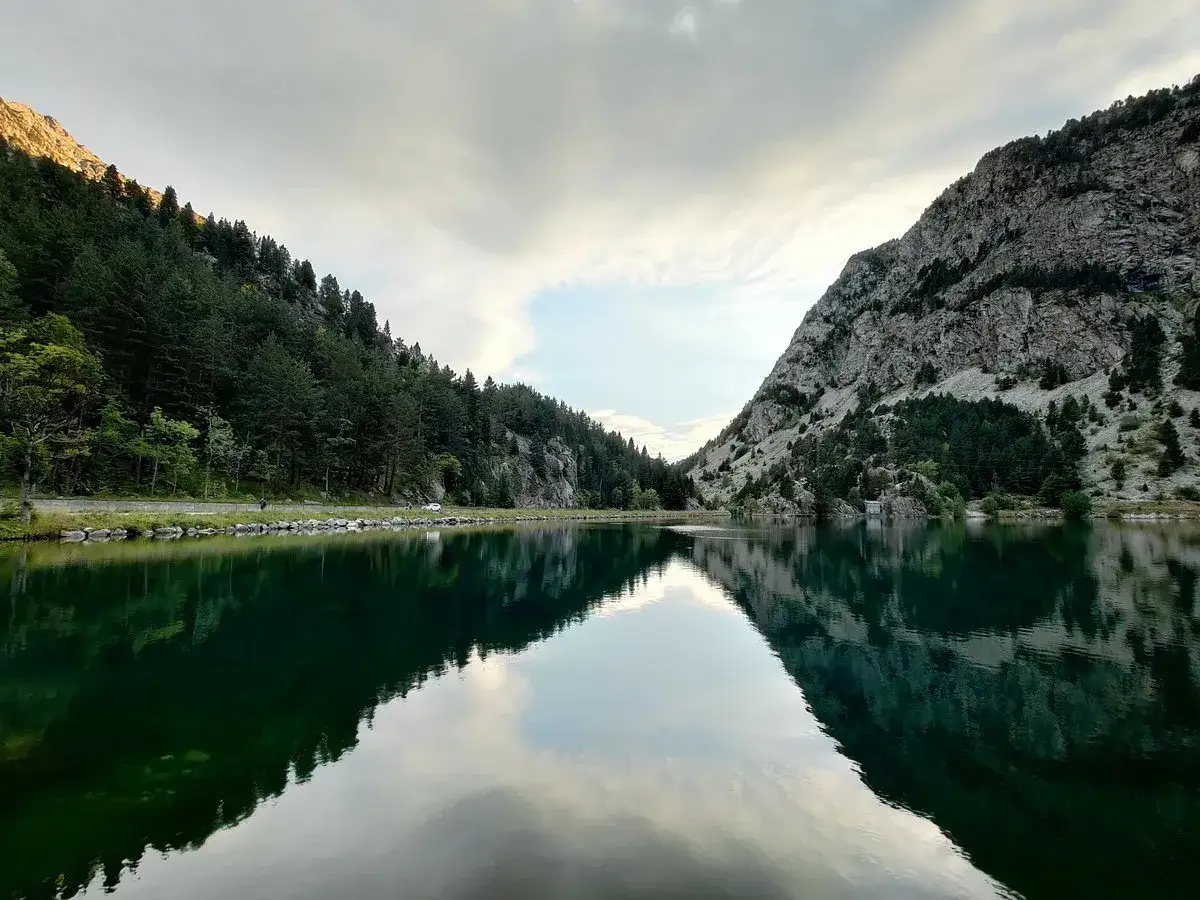 Reflejos perfectos en el ibon de Ba&ntilde;os, con monta&ntilde;as cubiertas de pinos y un cielo nublado.