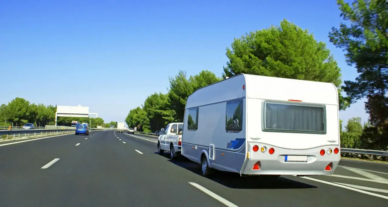 Un coche blanco remolca una caravana por una autopista soleada, rodeada de &aacute;rboles verdes. El transporte caravana promete aventuras.