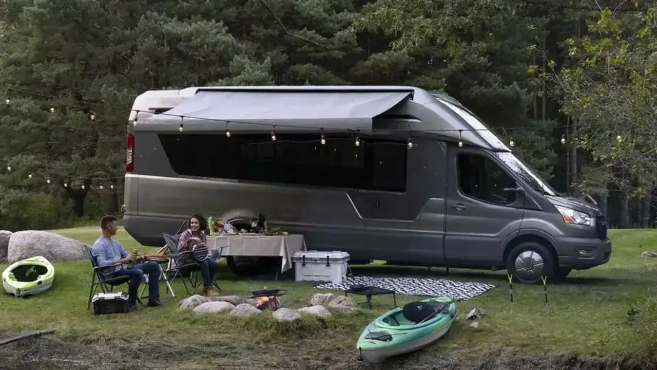 Pareja disfruta de una cena al aire libre junto a una vw california el&eacute;ctrica en un entorno boscoso, con luces y un kayak.