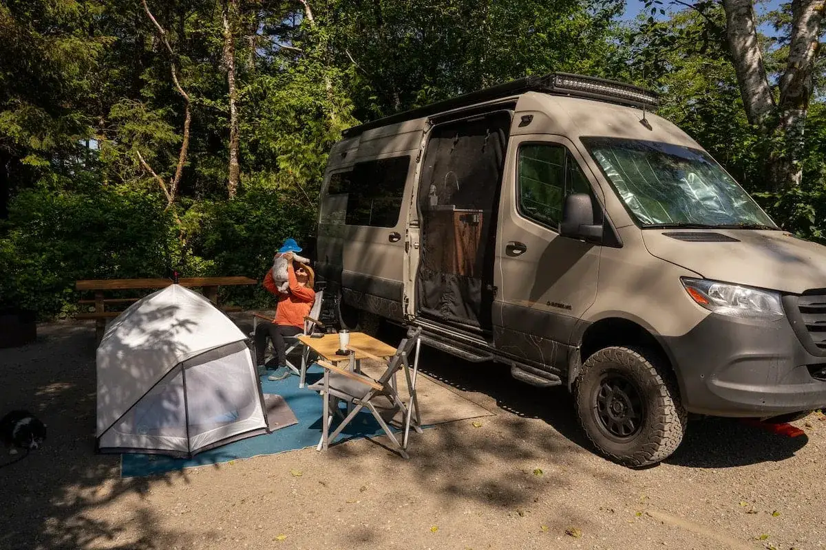 Una mujer con un gato en brazos se sienta junto a un veh&iacute;culo vivienda en un campamento. Hay una tienda de campa&ntilde;a y una mesa plegable.