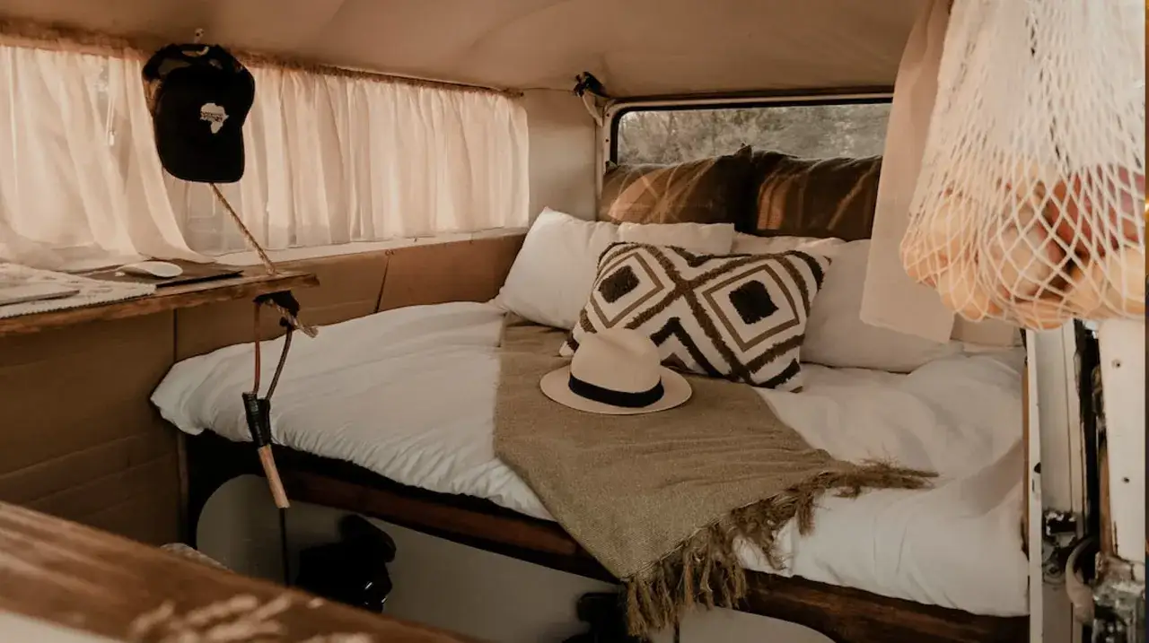 Interior de camper con cama, cojines, sombrero y una bolsa de malla con frutas. Ideas para como llevar la ropa en una camper de forma organizada.