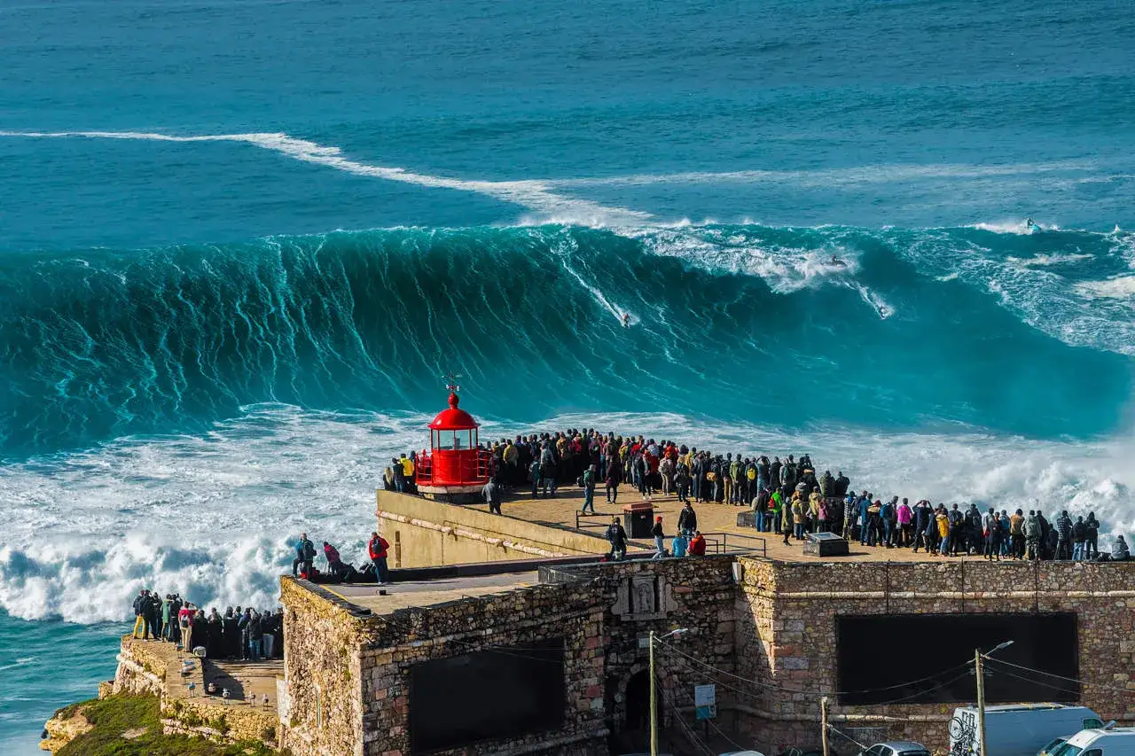Multitud observa las gigantescas olas de Nazar&eacute; desde un mirador con un faro rojo. Surfistas desaf&iacute;an la imponente fuerza del mar.