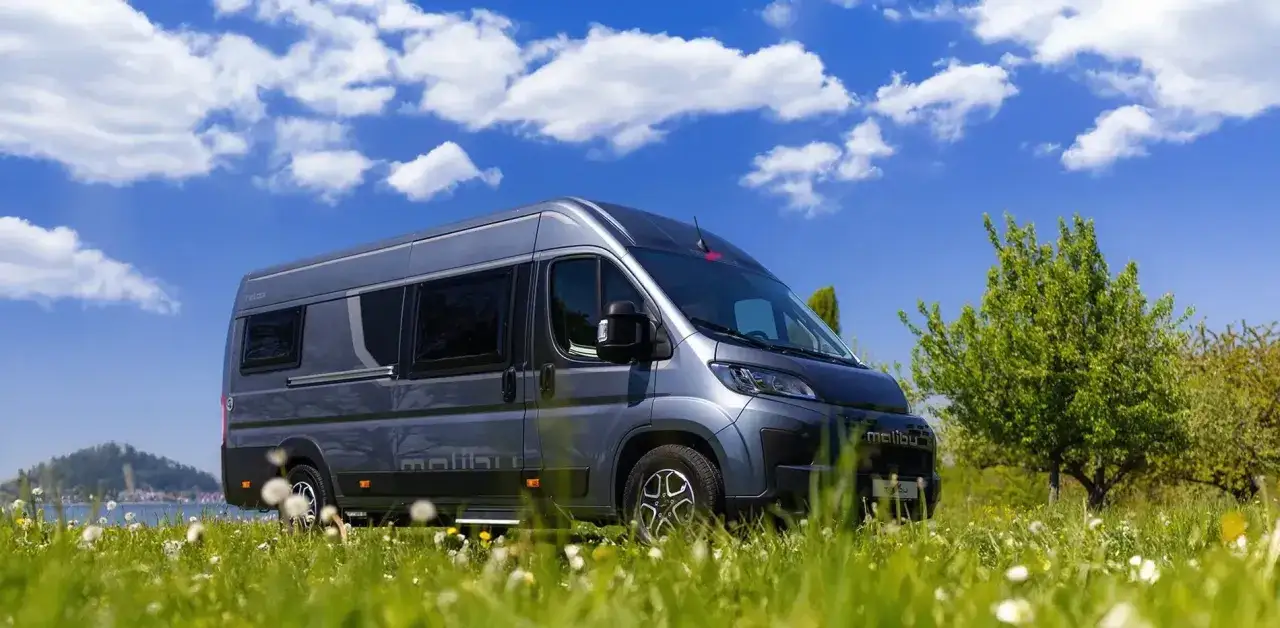 Autocaravana Malibu gris aparcada en un prado verde, con un lago y monta&ntilde;as al fondo. Ideal para explorar los concesionarios de autocaravanas en Alemania.