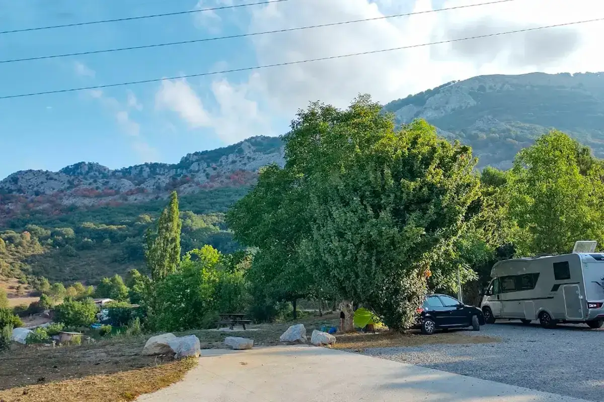 Un &aacute;rea camper en Pamplona con autocaravana y coche aparcados bajo &aacute;rboles frondosos, con monta&ntilde;as de fondo.