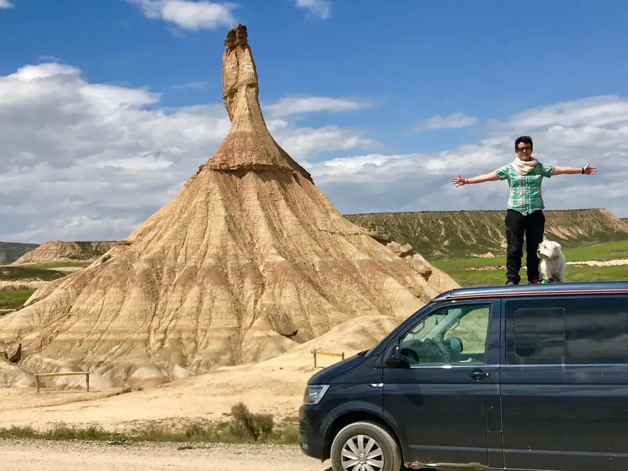 Aventuras en Navarra en camper: persona y perro en el techo de la furgoneta, con un paisaje des&eacute;rtico y una formaci&oacute;n rocosa imponente.