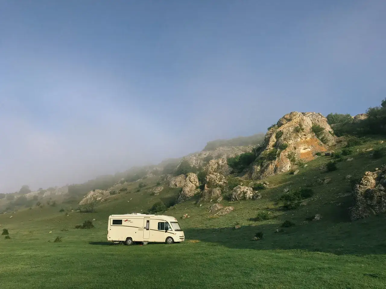 Una camper aparcada en un prado verde, con una monta&ntilde;a rocosa y niebla al fondo. Aventura transpirenaica en camper.