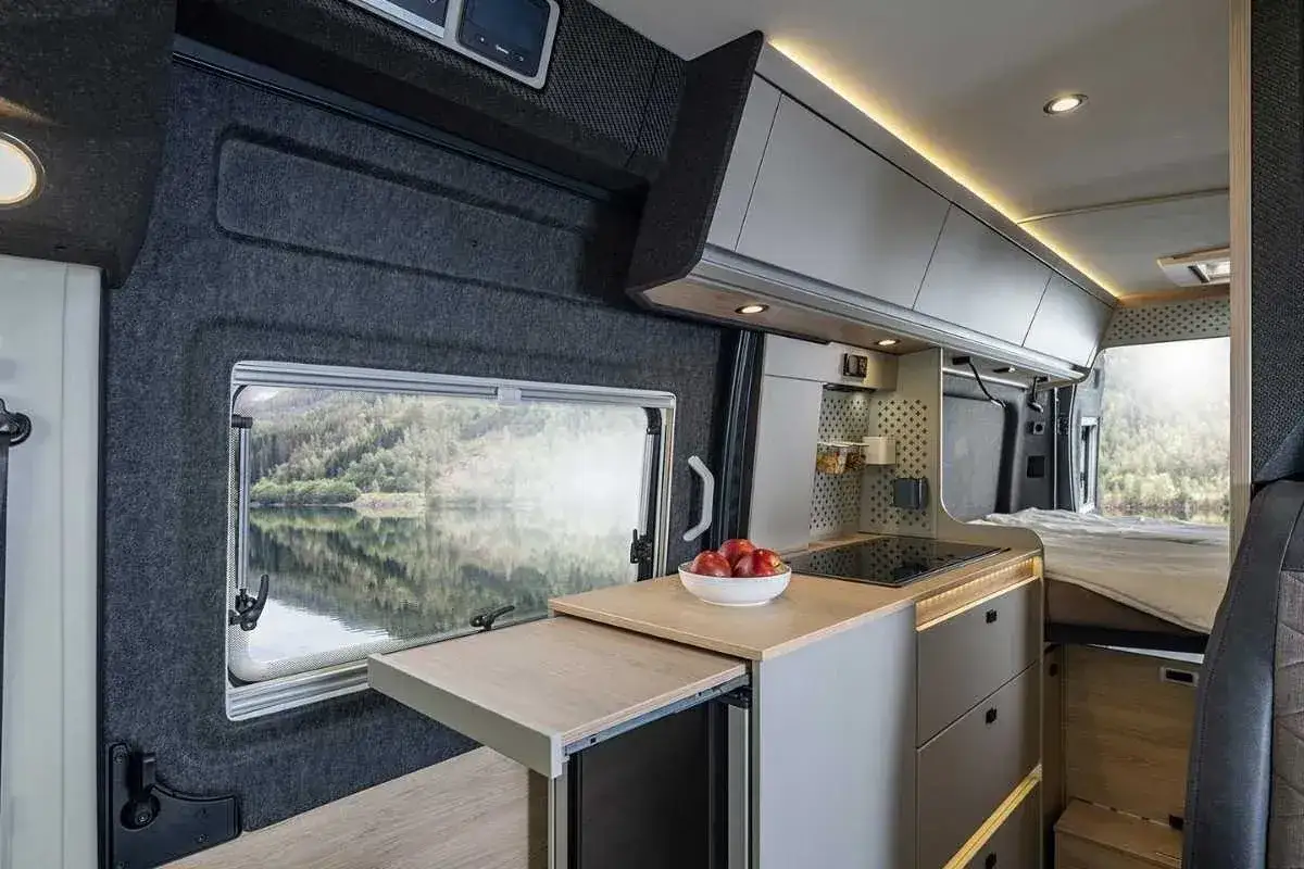 Cocina de un camper garage con vista a un lago. Muebles modernos, encimera con vitrocer&aacute;mica y cama visible.