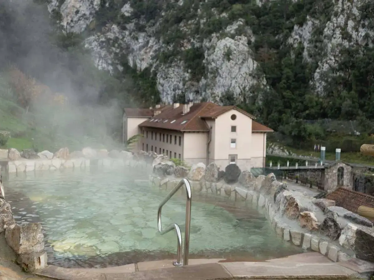 Piscina de aguas termales en Espa&ntilde;a, rodeada de rocas y con vapor ascendiendo. Al fondo, un edificio y monta&ntilde;as verdes.