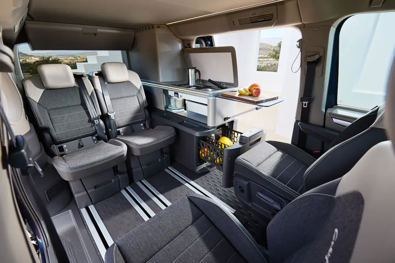 Interior de una VW California con asientos cómodos, cocina compacta y cesta de frutas. ¡Lista para la aventura!