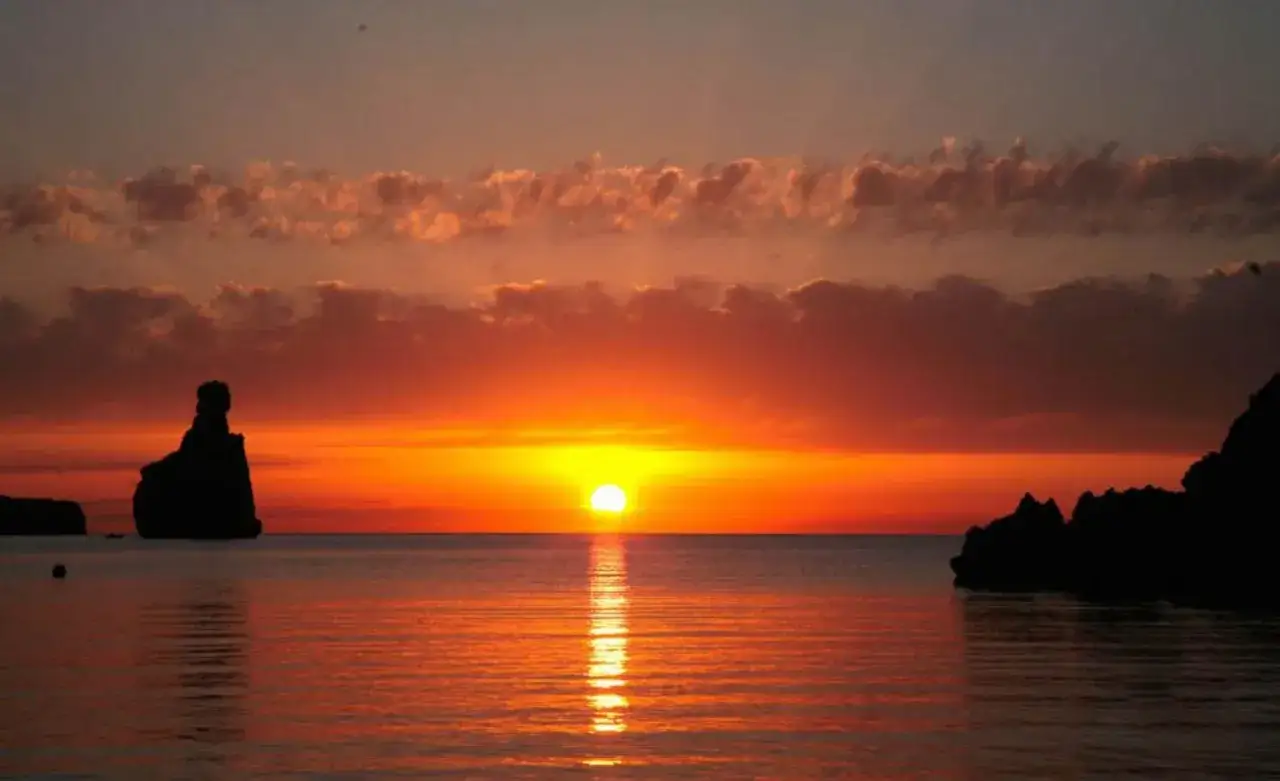Espectacular benirras tramonto con el sol ti&ntilde;endo el cielo de naranja y reflej&aacute;ndose en el mar. Siluetas rocosas emergen del agua.