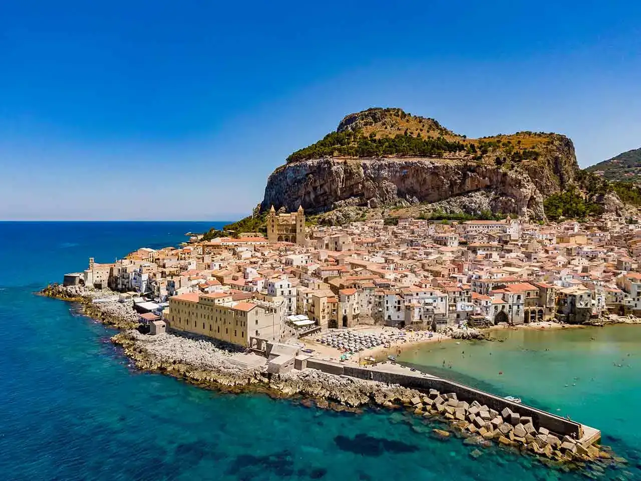 Vista a&eacute;rea de Cefal&uacute;, Italia, con sus casas blancas y tejados naranjas extendi&eacute;ndose hasta el mar azul turquesa, bajo un imponente pe&ntilde;&oacute;n.