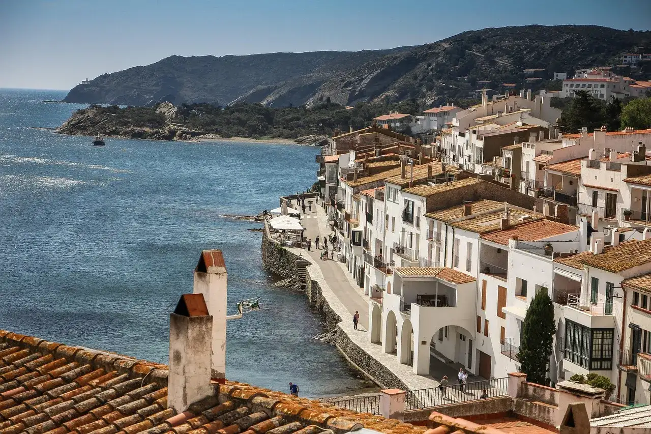 Pueblo costero blanco en Catalu&ntilde;a, ideal para &aacute;reas camper. Mar azul, casas con tejados naranjas y colinas verdes.