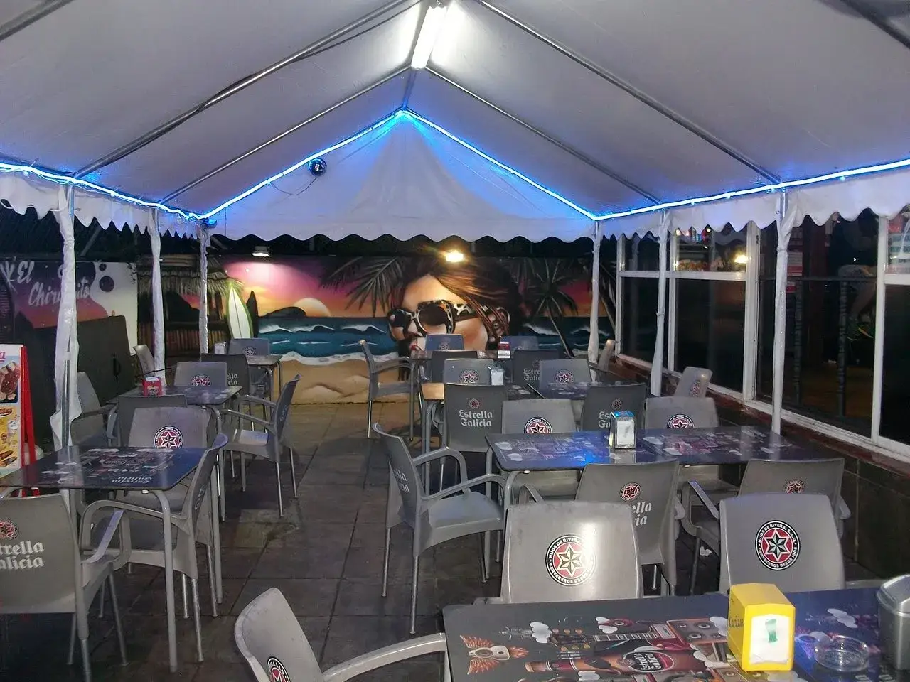 Terraza de un chiringuito con mesas y sillas listas para comer en Ori&ntilde;&oacute;n. Mural playero de fondo.
