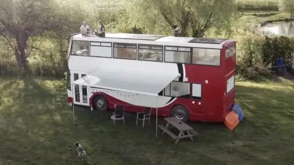 Autobús motorhome de dos pisos con paneles solares en el techo. Personas sentadas en la parte superior, un toldo extendido y muebles de exterior.