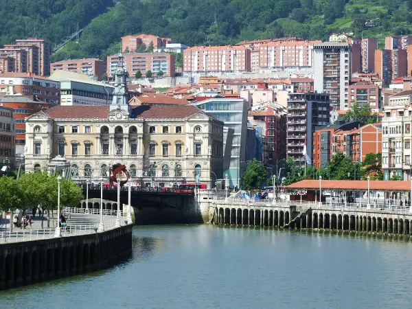 Vista del río Nervión y el Ayuntamiento, un edificio emblemático del bilbao centro storico, con edificios modernos y colinas verdes al fondo.