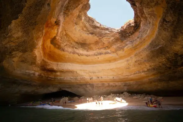 Interior de una cueva marina con un agujero en el techo, donde está Benagil. Gente en kayaks y en la playa disfrutan del sol.
