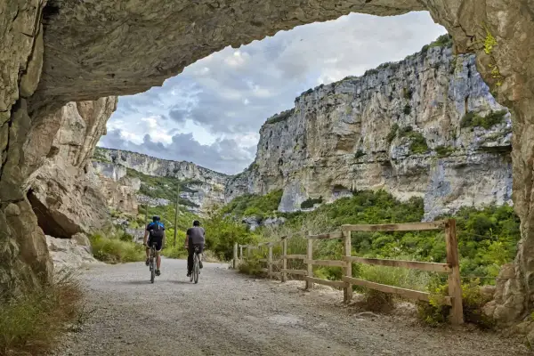 Dos ciclistas exploran la impresionante **foz de Lumbier**, un cañón rocoso con vegetación exuberante y un cielo nublado.