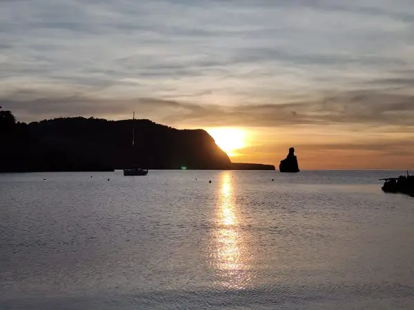 Un velero navega en la serena bahía mientras el sol se pone en Benirràs, tiñendo el cielo de tonos dorados y naranjas.