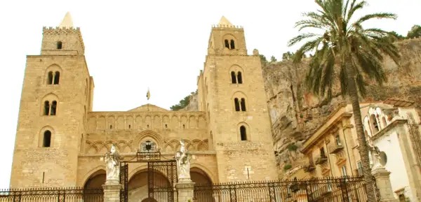 La imponente catedral de Cefalú se alza majestuosa contra un acantilado rocoso, con una palmera que añade un toque tropical a la escena.